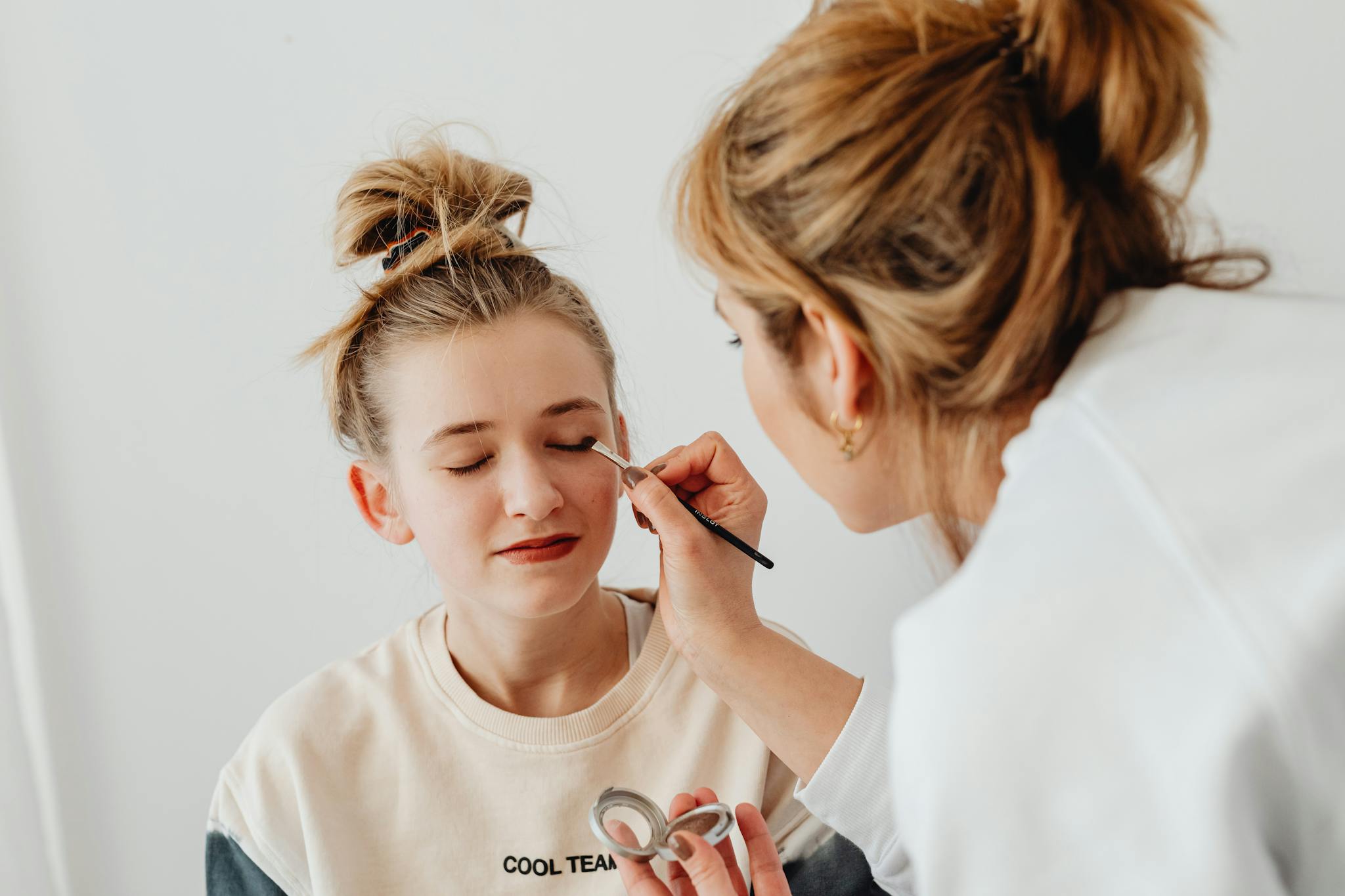A makeup artist skillfully applies eyeshadow on a teenager with a relaxed expression.