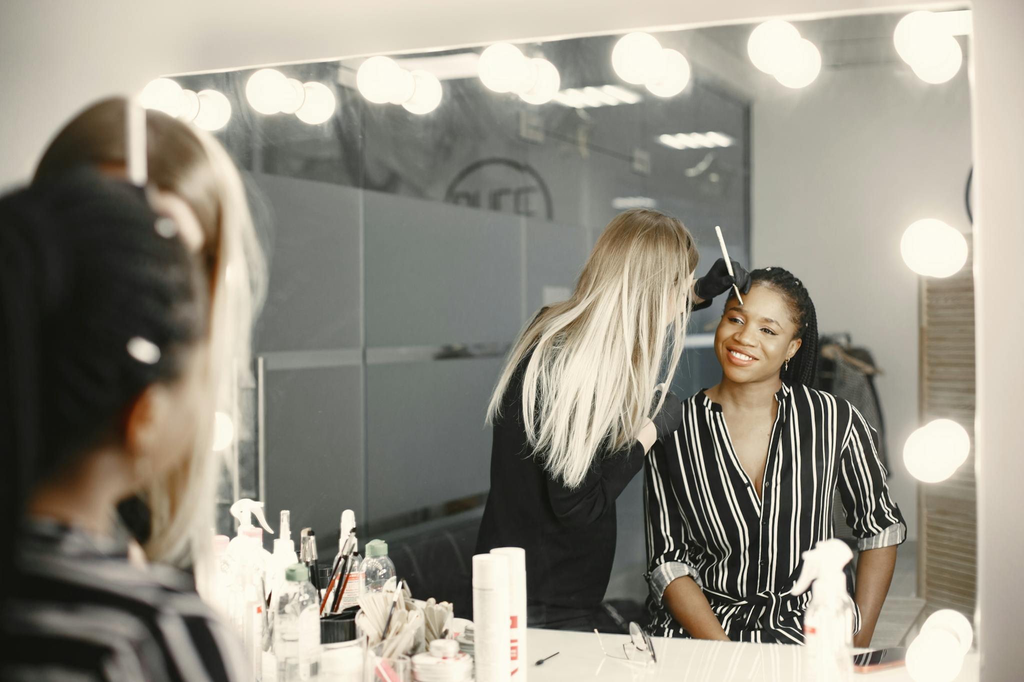 A professional makeup artist applies makeup to a smiling woman in a well-lit backstage setting.